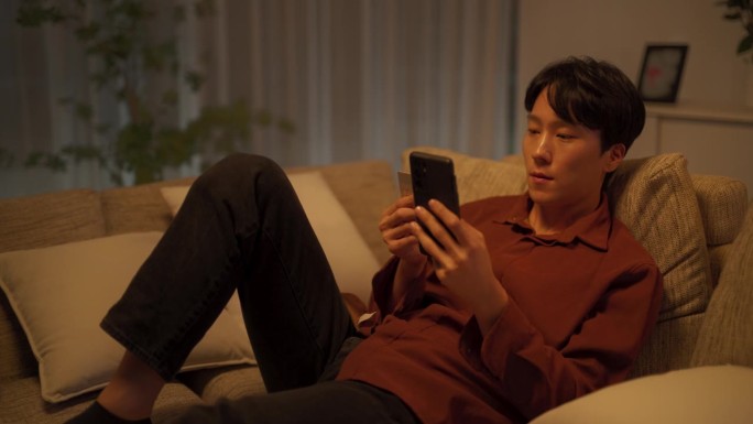 一名年轻的韩国男子在舒适的公寓里使用网上购物App，用智能手机和信用卡安全地购物，为自己购买最新的时