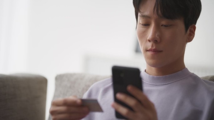 一个时尚的韩国男人坐在舒适的客厅的沙发上，用智能手机网购，拥抱移动电子商务解决方案的便利