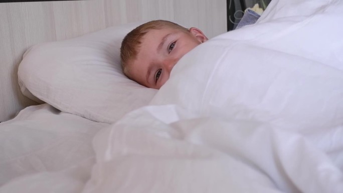 一个可爱的八岁男孩不想睡觉，沉迷于白色亚麻布的床上。