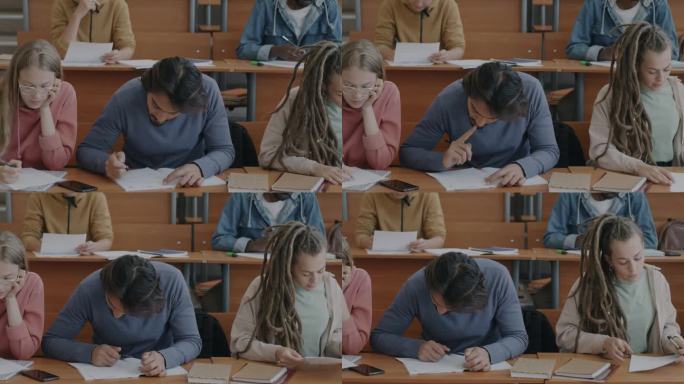 多民族的男女学生坐在礼堂的桌子旁写考试试卷
