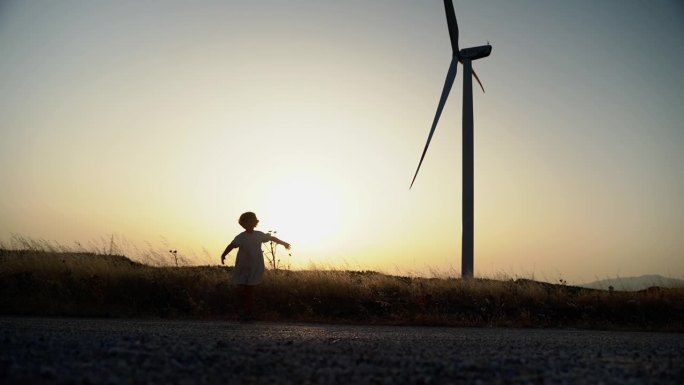 快乐的孩子女孩的剪影旋转，转身双手在大自然的夕阳在田间与风车风力发电机。自由，童年，环保能源动力。
