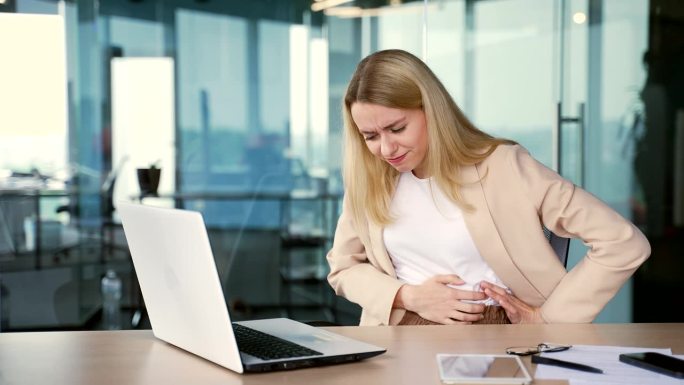 患有胃痛的年轻女商人坐在现代办公室的工作场所用笔记本电脑工作
