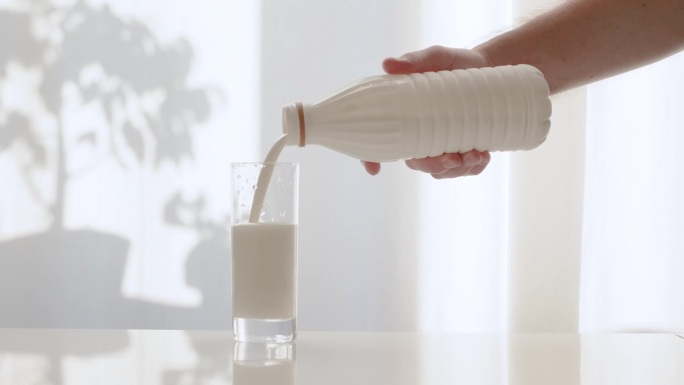 把牛奶从塑料瓶里倒进玻璃杯里。牛奶在白色天鹅绒窗帘的背景下倾泻而下。厨房桌子上的透明玻璃杯里装着新鲜