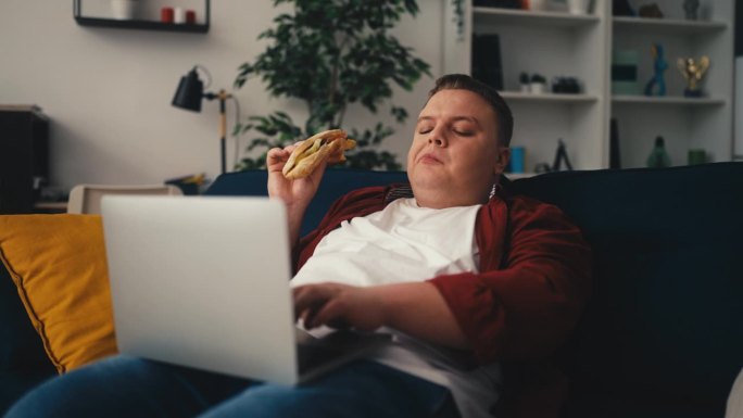 肥胖男性使用笔记本电脑，在沙发上吃三明治，久坐不动的生活方式