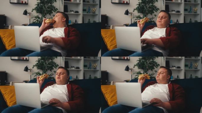 肥胖男性使用笔记本电脑，在沙发上吃三明治，久坐不动的生活方式
