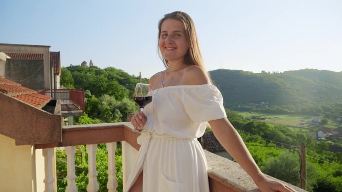 面带微笑的黑发女子，端着一杯红酒，站在山间别墅的阳台上，映衬着令人惊叹的日落。旅行，夏日，享受大自然