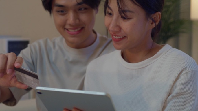 年轻的亚洲夫妇玩数码平板设备购物，使用信用卡在线支付一起坐在沙发上在家里的客厅晚上。生活方式花时间在