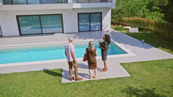 女房地产经纪人向一对站在后院的夫妇展示一栋带游泳池的现代住宅
