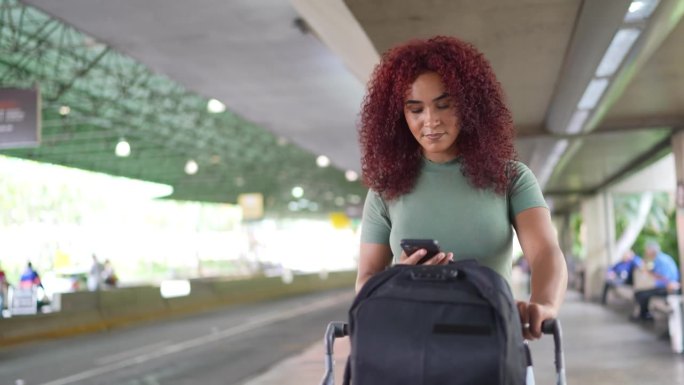 一名年轻女子在机场边走边推行李车边玩手机