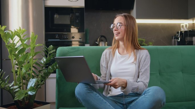 年轻漂亮的20岁女孩，戴着眼镜，像自由职业者一样在笔记本电脑上打字，而坐在家里的厨房背景里。呆在家里