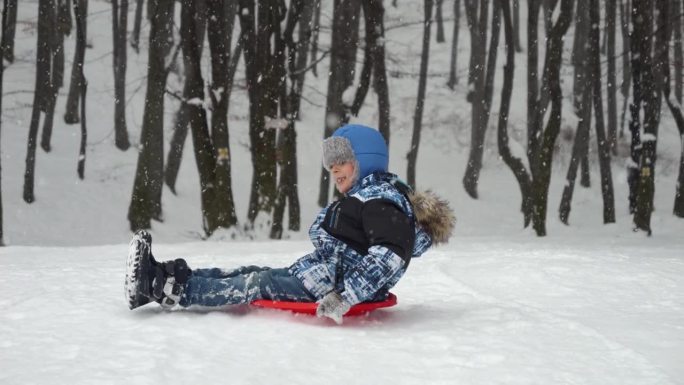 一个快乐的小男孩骑着红色的塑料雪橇从雪坡上滑下来，他的脸上洋溢着兴奋和笑声。冬天玩耍的时光和无拘无束