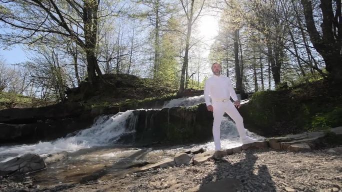 一个穿白衣的男人在森林里唱歌，背景是瀑布和自然