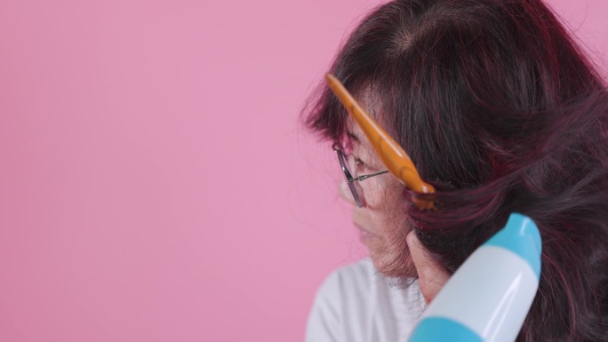 老妇人用吹风机在粉红色的背景上吹干她的头发