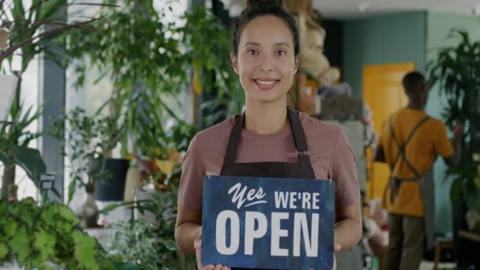 美丽的中东女士的肖像，系着围裙，站在花店里，手里举着“Yes, we are open”的牌子