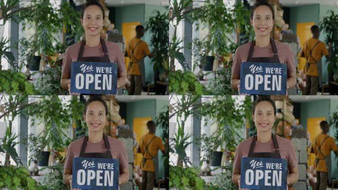 美丽的中东女士的肖像，系着围裙，站在花店里，手里举着“Yes, we are open”的牌子