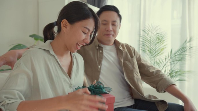 快乐的亚洲夫妇坐在家里客厅的沙发上，用红色礼盒送生日礼物，玩得很开心。生活方式花时间在家里。