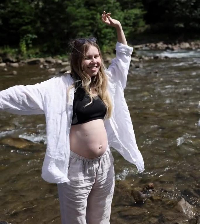 一位身穿白色连衣裙的孕妇站在山间河流的水中，挥动着双手，连衣裙的布料在风中飘动。垂直视频
