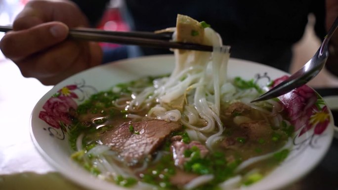 一名男子在金属桌子上用筷子和勺子吃着越南传统的Pho Bo汤、牛肉和米粉