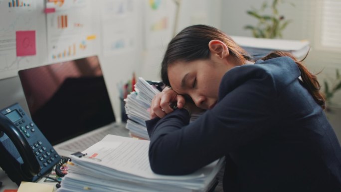 疲倦困倦的年轻女商人在办公桌上堆着一叠文件睡着了