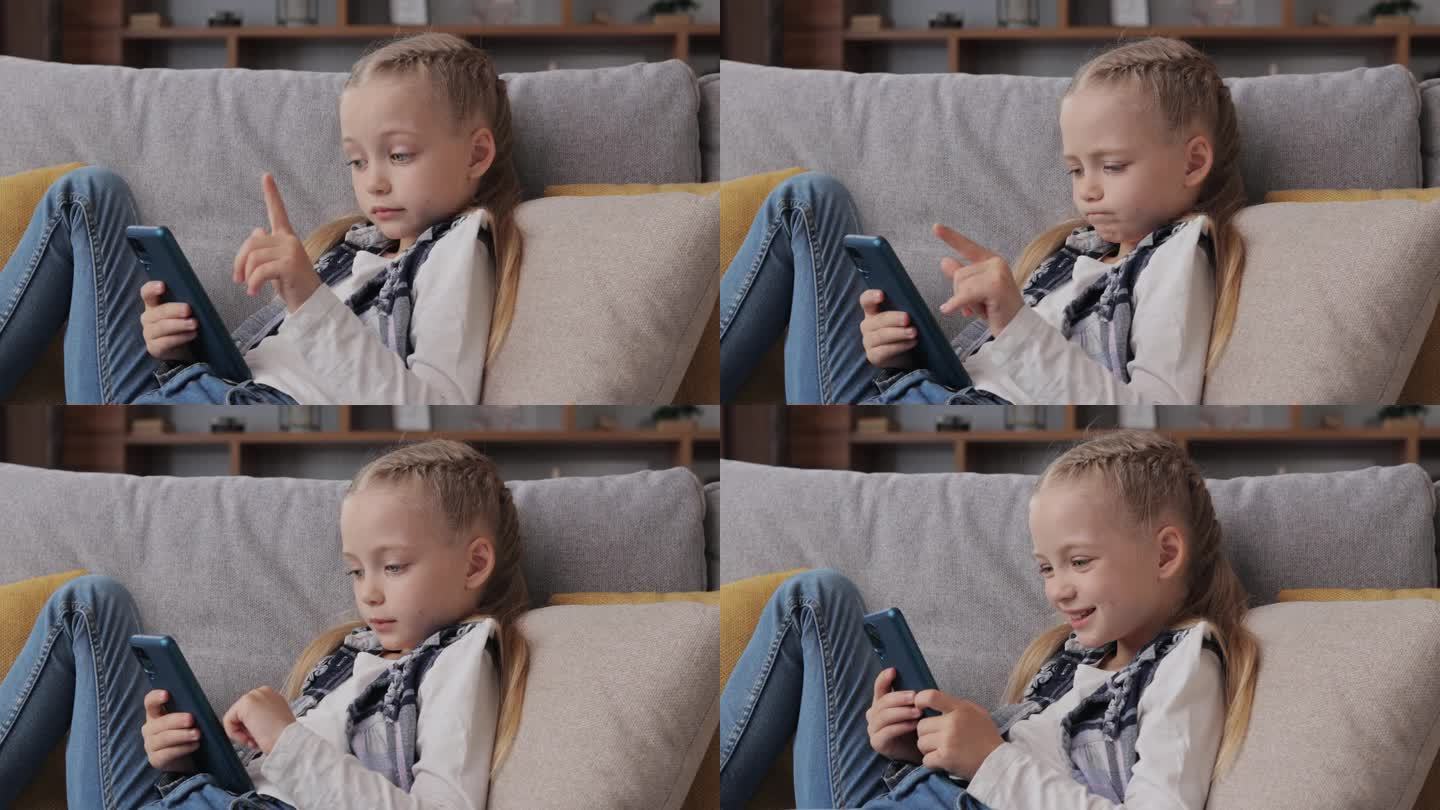 惊讶的可爱小女孩抱着智能手机躺在家里的沙发上。选择最喜欢的音乐或动画片，发短信，上网，看视频，玩智能