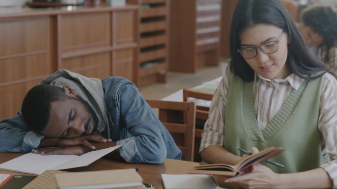 亚洲女人在大学图书馆学习，而疲惫的非洲裔美国男人懒惰的学生睡觉