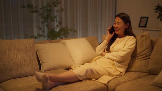 晚上，在一间舒适的公寓里，一个美丽的韩国女人躺在沙发上，微笑着与朋友进行有趣的电话交谈，分享关于关系