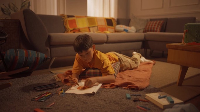 年轻的韩国男孩躺在地板上，用钢笔、铅笔、水彩画在笔记本上，在温暖的光线下舒适的公寓里。未来的艺术家是