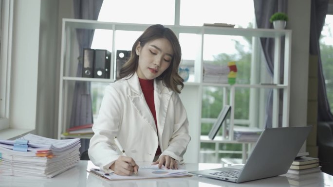 有魅力的亚洲女商人使用笔记本电脑，检查桌上的财务文件，计算财务报告。用笔记本电脑和计算器整理文件。