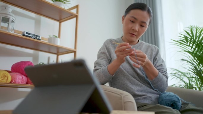 亚洲妇女坐在家里的客厅里，从平板电脑上的在线视频剪辑中学习如何钩针编织。