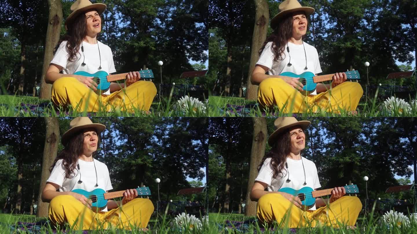 公园里的女孩坐在草地上弹尤克里里。学习演奏四弦乐器。在公园里放松。音乐疗法，心理学，冥想，放松，放松