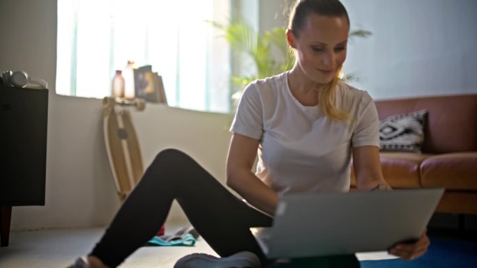慢动作肖像微笑，美丽的年轻女子使用笔记本电脑，准备在线家庭锻炼在客厅的地板上。健康的生活方式，健身，