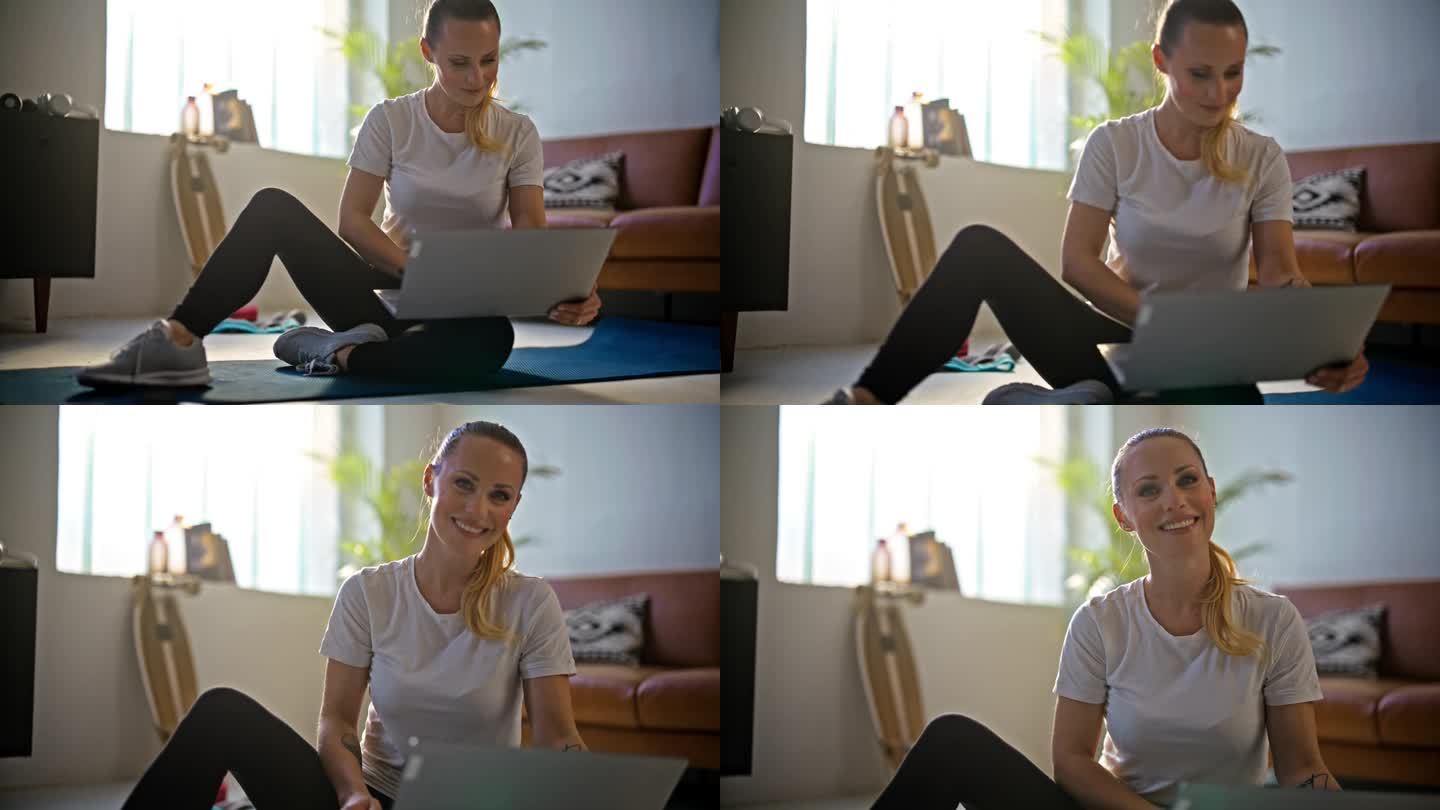 慢动作肖像微笑，美丽的年轻女子使用笔记本电脑，准备在线家庭锻炼在客厅的地板上。健康的生活方式，健身，