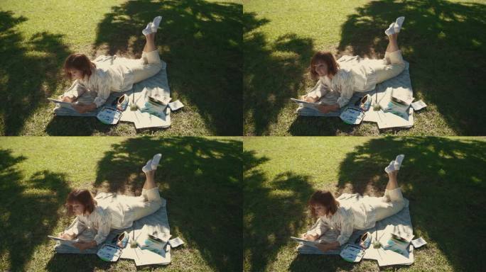 一个成年妇女躺在绿色公园的毯子上，在速写本上画画