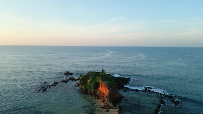 绿色的热带岩石岛在蓝绿色的海洋中间，在日落时被海浪冲刷。无人机拍摄的日出画面显示，斯里兰卡米瑞萨的鹦