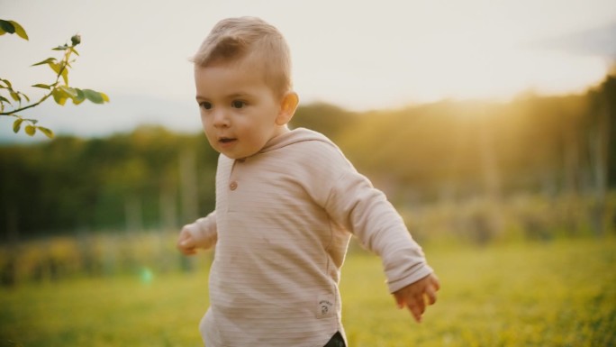 慢镜头:小男孩走在草地的斜坡上。可爱的小男孩喜欢在户外草地上散步，时而跌倒，时而爬起来。15个月大的