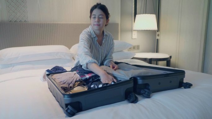 准备旅行，亚洲妇女穿着家居服收拾旅行箱准备旅行，坐在家里的床上。