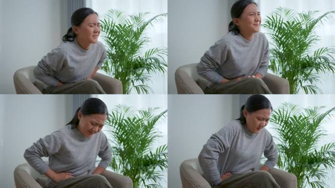 一位亚洲妇女胃痛，坐在家里客厅的扶手椅上。