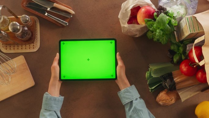 俯视图的匿名女性使用数字平板电脑与模拟绿色屏幕Chromakey显示在厨房的桌子上。女人在准备晚餐时