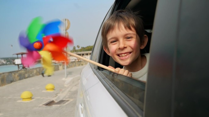 微笑的男孩看着打开的车窗，手里拿着彩色的纺车，因为风吹。儿童旅游、旅行、旅游、假期、周末和假期的概念