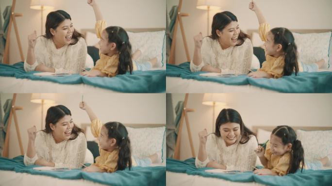 亚洲母亲在家教女儿做作业，在床上玩耍。年轻的母亲在家照顾女儿并教她画画。