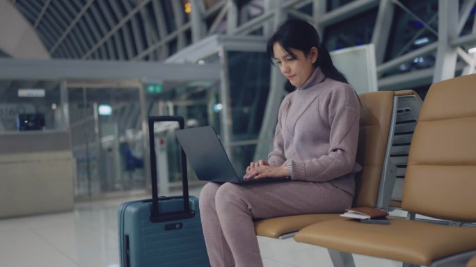 一名紫衣女子拎着行李箱和护照独自坐在机场，在登机前使用笔记本电脑工作