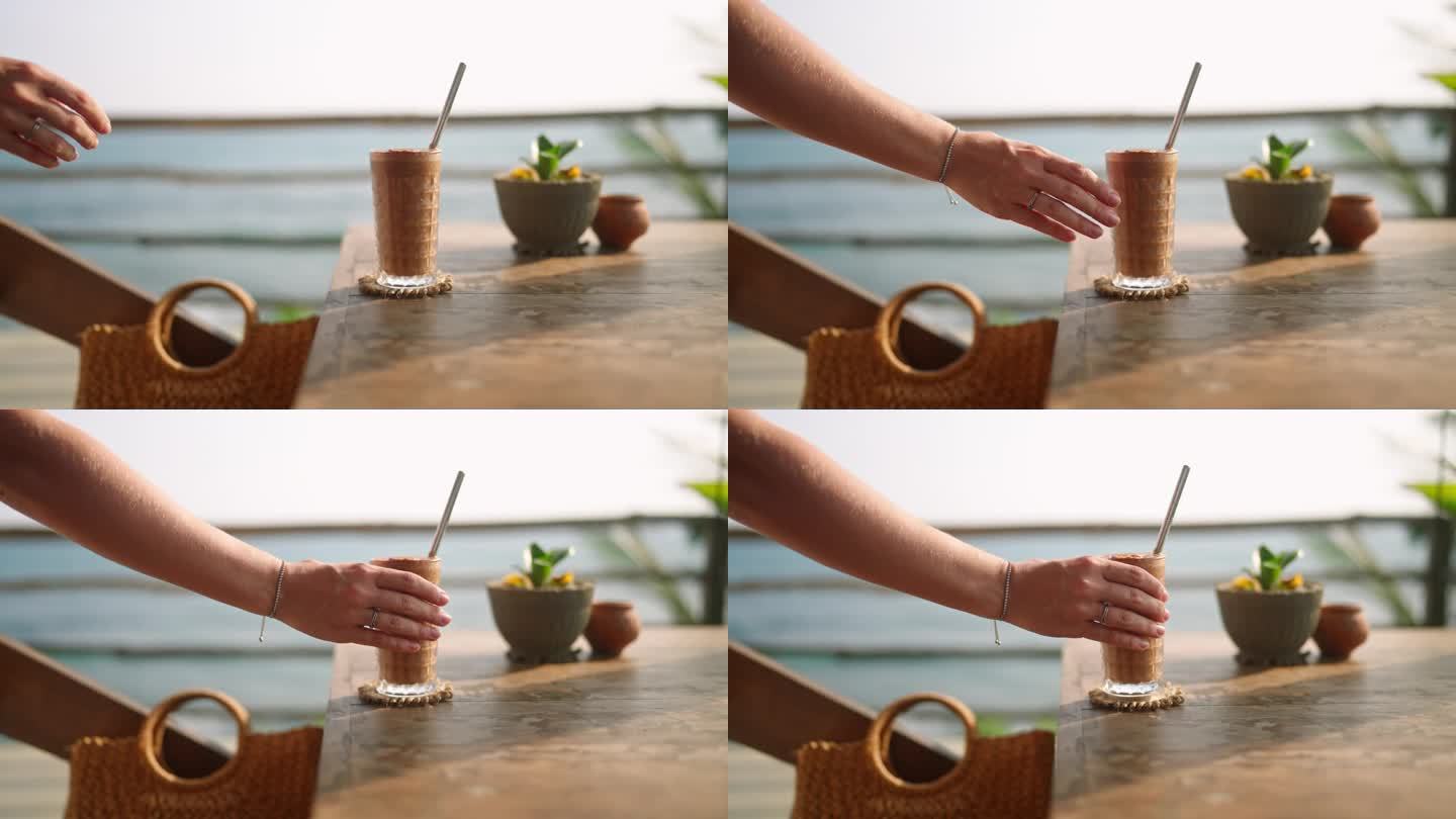 在室外热带海景咖啡馆，一位女士从木桌上拿起装有鸡尾酒和金属吸管的高脚杯。将冰咖啡或冰沙放到观景屋顶餐