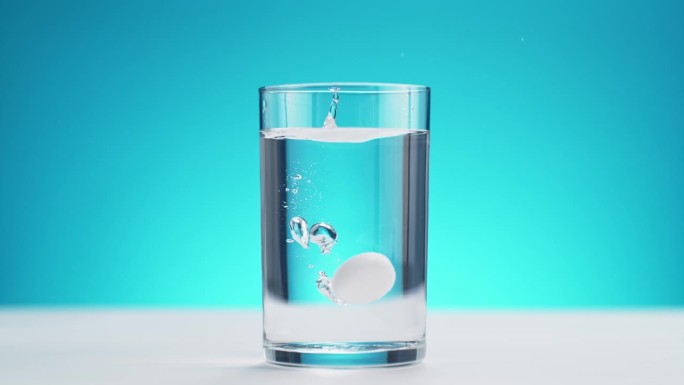 白色桌面上的泡腾片落入玻璃杯中，蓝色背景下的慢动作溅起水花