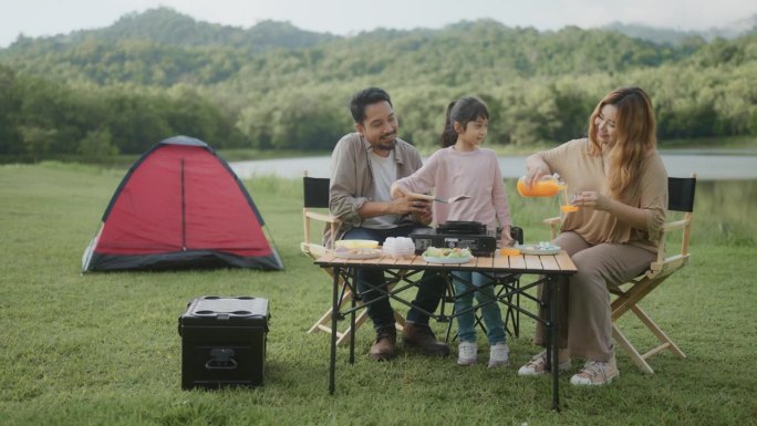一个亚洲家庭带着女儿一起去露营，女儿在帐篷里为家人做早餐。
