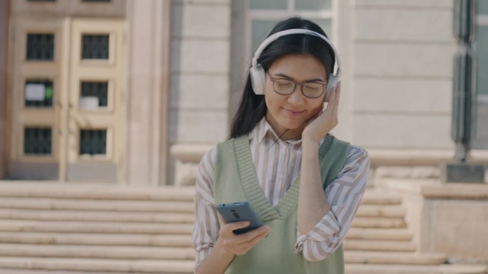 一个快乐的亚洲女人，戴着耳机，拿着智能手机，听着音乐，走在城市里