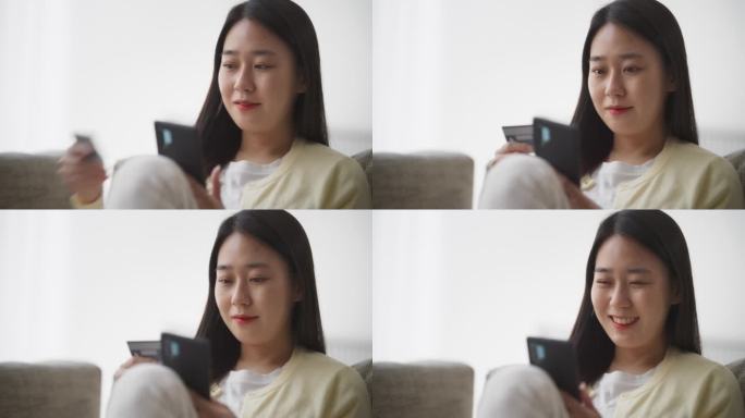 一个漂亮的韩国女性用智能手机拿着信用卡的肖像。亚洲女性做移动购物疗法，在客厅浏览网上购物网站，发现时