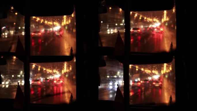 从小巴的挡风玻璃内看阴雨天气下的夜晚城市和开着前灯的汽车。雨滴落在玻璃上。