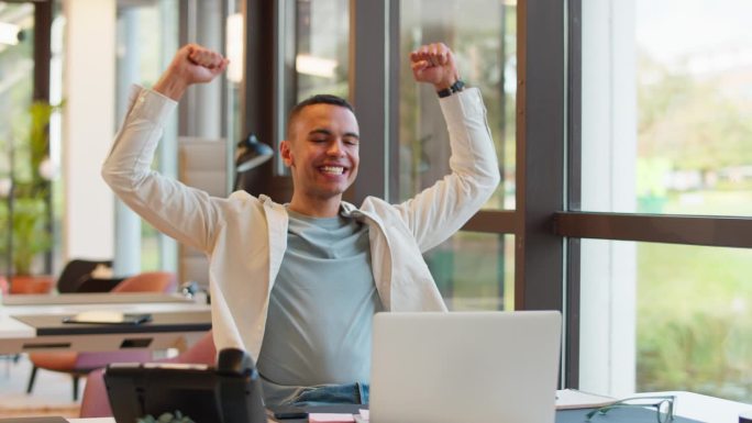 兴奋的年轻商人拿着笔记本电脑庆祝好消息在办公室办公桌前工作