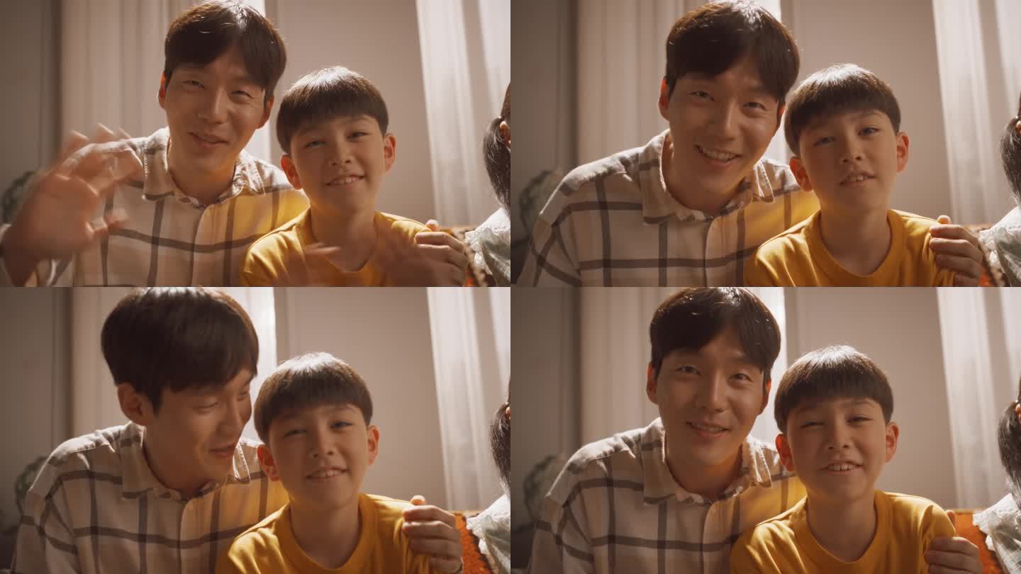幸福的韩国父子在家里的客厅里，微笑着对着镜头挥手。屏幕更换与朋友在线视频通话。家庭使用科技保持联系