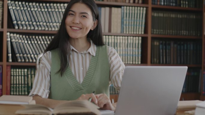 无忧无虑的亚洲女大学生的肖像，坐在图书馆的书桌前，微笑地看着镜头